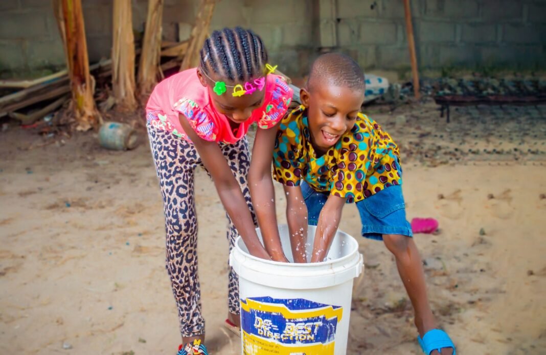 Two smiling African children washing their hands together in a bucket of water outdoors