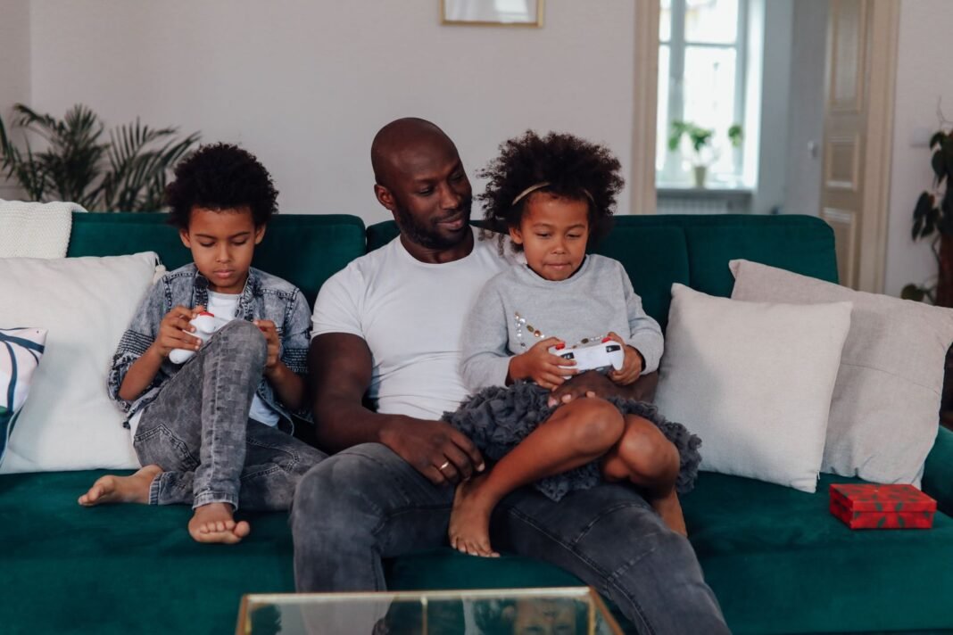 Father sitting on the sofa with his son and daughter while they play video games with controllers
