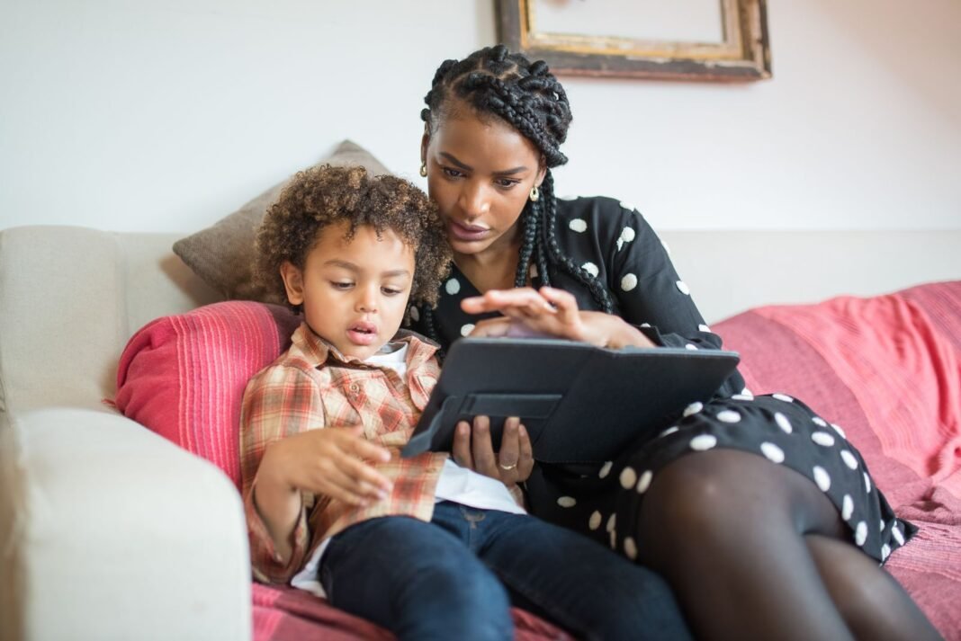 Mother sitting on a sofa with her young son, guiding him as they use a tablet together